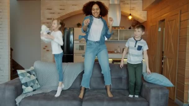 Wanita Afrika Amerika dan anak-anak Kaukasia melompat di sofa di rumah bersenang-senang menari — Stok Video