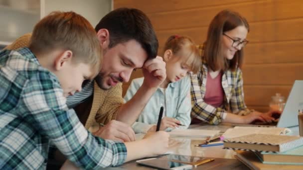 Щасливий хлопець робить домашнє завдання з маленьким сином, розмовляючи сміється, коли жінка навчає доньку вдома — стокове відео