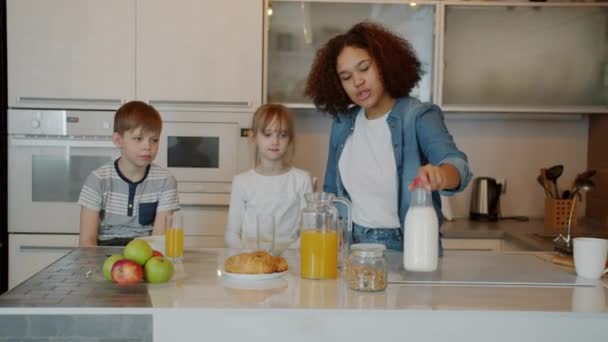 A gondoskodó bébiszitter tejet tölt a fiúknak és a lányoknak, eteti a gyerekeket otthon a konyhában. — Stock videók
