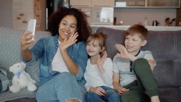 Babysitter amichevole e simpatici bambini che parlano e salutano mano durante la videochiamata online su smartphone — Video Stock