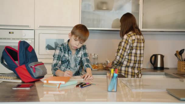 Skolpojke gör läxor medan vårda mamma matlagning mat i köket — Stockvideo