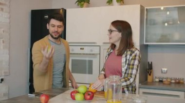Neşeli genç adam elmayla hokkabazlık yaparken hamile kadın evde yemek pişirirken gülüyor.