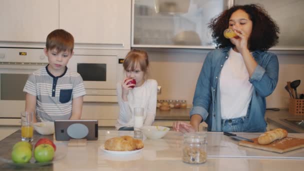 Niña y niñera viendo divertido espectáculo en la pantalla de la tableta riendo durante el desayuno en la cocina — Vídeos de Stock