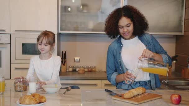 Маленькая девочка наполняет тарелку хлопьями, пока няня наливает сок в стакан дома на кухне — стоковое видео