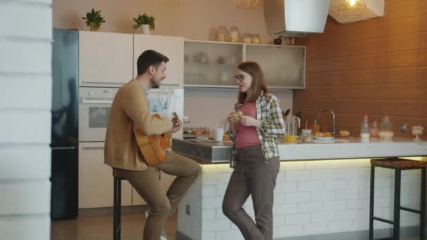 Jongeman speelt gitaar terwijl zwangere vrouw fruit eet luisteren naar muziek en glimlachen in de keuken thuis — Stockvideo
