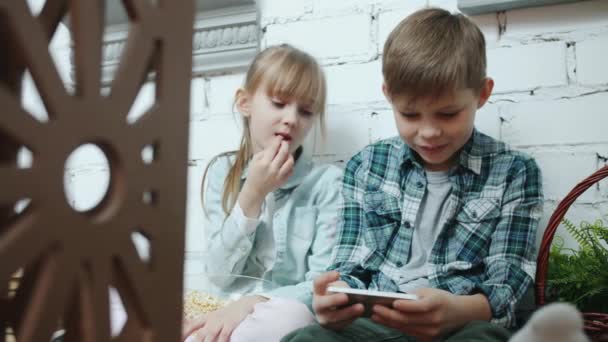 スマートフォンでビデオゲームをしているかわいい子供たちのスローモーションポップコーンを食べて笑う — ストック動画
