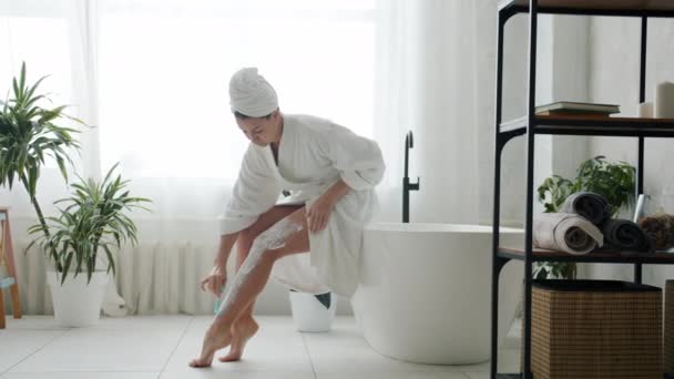 Mooie jonge vrouw in badjas scheren benen zorgdragen voor lichaam in badkamer — Stockvideo