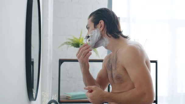 Movimiento lento de chico cerda de afeitar en la cara con maquinilla de afeitar moderna mirando el espejo en el baño — Vídeo de stock