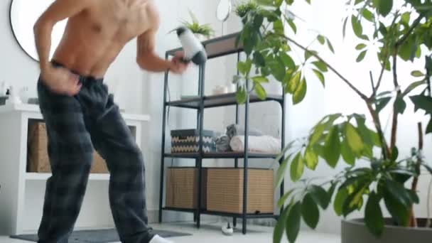 Portret van zorgeloze man die plezier heeft in de badkamer dansen en zingen holding föhn — Stockvideo