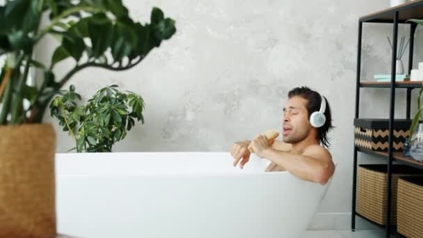 Змішана раса чоловік танцює і співає в пензлі, насолоджуючись музикою в навушниках, приймаючи ванну вдома — стокове відео
