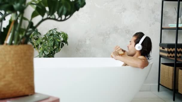 Ελκυστικό νεαρό μελαχρινό τραγουδώντας σε βούρτσα μαλλιών ακούγοντας μουσική με ακουστικά στην μπανιέρα — Αρχείο Βίντεο