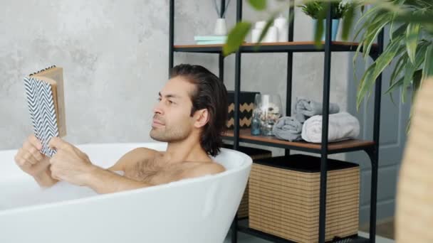 Hombre inteligente de raza mixta disfrutando de libro leyendo historia en bañera relajante y estilizando en baño — Vídeo de stock