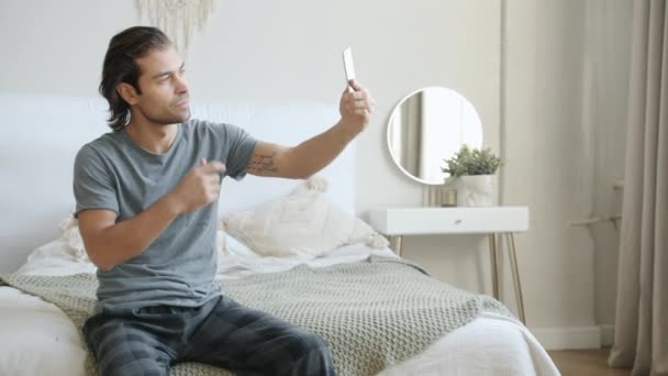 Гарний молодий чоловік бере селфі, використовуючи камеру смартфона, сидячи в ліжку вдома — стокове відео