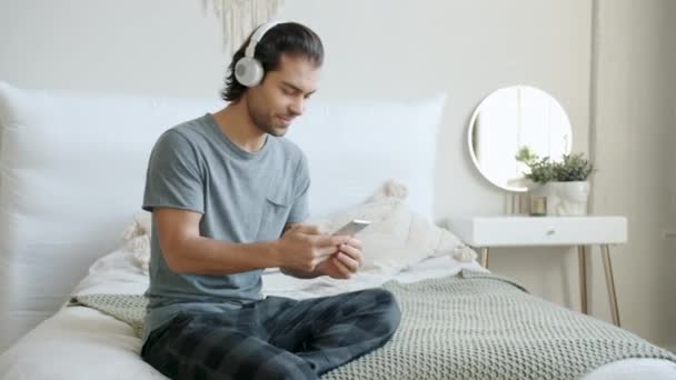 Hombre de raza mixta escuchando música en auriculares usando teléfonos inteligentes en el interior de la cama — Vídeos de Stock