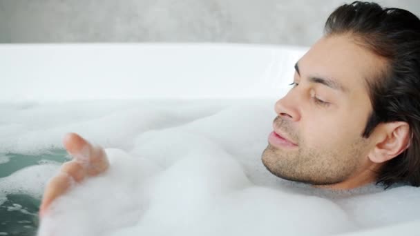 Close-up de cara despreocupado relaxando na banheira e se divertindo com espuma de sabão no banheiro — Vídeo de Stock