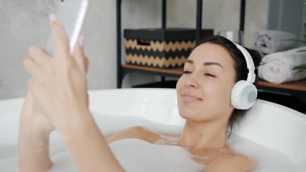 Långsam rörelse av avslappnad flicka i hörlurar njuter musik dans med smartphone i badkaret — Stockvideo