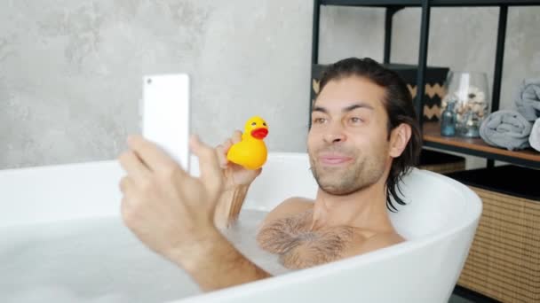 Cámara lenta de tipo alegre tomando selfie con pato de juguete tomando baño con teléfono inteligente — Vídeos de Stock