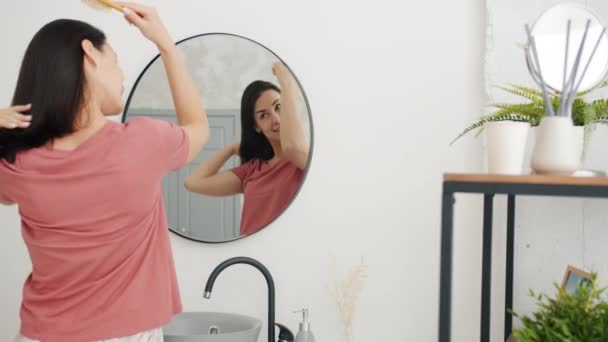 Mouvement lent de jolie étudiante brossant les cheveux en regardant miroir de salle de bains — Video