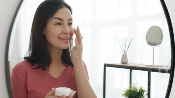 Ευτυχισμένη νεαρή γυναίκα που βάζει κρέμα στο δέρμα του προσώπου χαμογελώντας και κοιτάζοντας τον καθρέφτη στο μπάνιο — Αρχείο Βίντεο