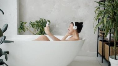 Kulaklık takan, küvette müzik dinleyen, dans eden ve akıllı telefon kullanan neşeli bir kadın.