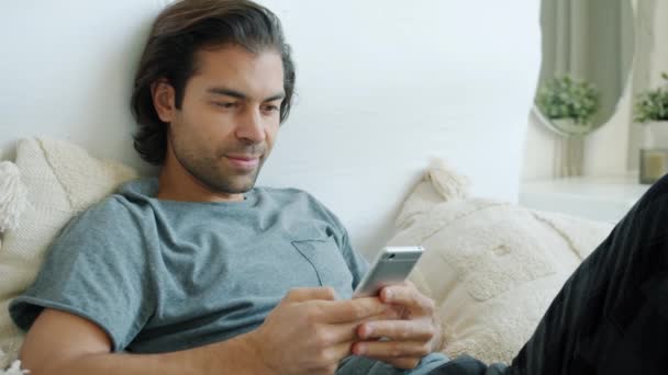 Веселий змішана гонка хлопець прочищає екран смартфона, насолоджуючись сміхом у соціальних мережах, розважаючись у ліжку — стокове відео