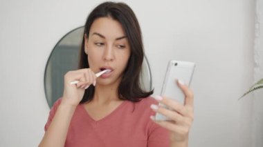 Ciddi bir kadın dişlerini fırçalıyor ve banyodaki ekrana bakarak modern akıllı telefon kullanıyor.