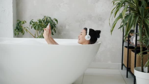 Jovem atraente relaxando na banheira usando smartphone e ouvindo música com fones de ouvido — Vídeo de Stock