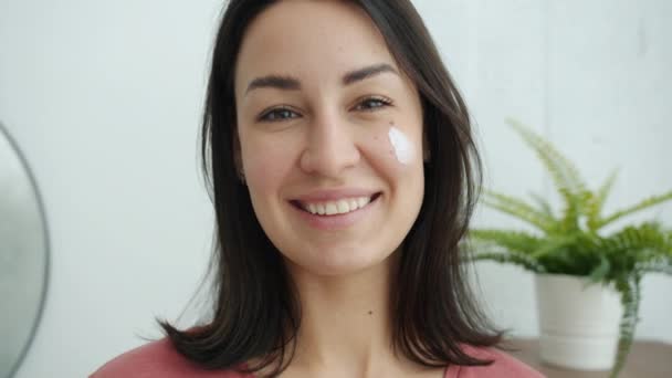 Portrait of pretty woman with cream on face smiling looking at camera holding cosmetic product — Stock Video