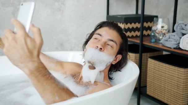 Chico feliz tomando selfie divertido con barba de espuma usando teléfono inteligente en la bañera — Vídeos de Stock