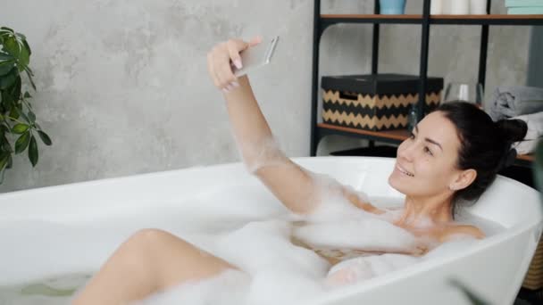 Радостная молодая женщина позирует перед камерой со смешными лицами, делая селфи в ванной дома — стоковое видео