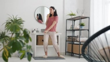 Evde banyoda, saç fırçasında şarkı söyleyen, kulaklık takan mutlu genç bir kadın.