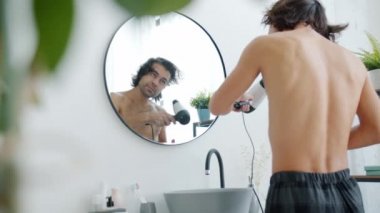 Pijamalı, ıslak, uzun saçlı, saç kurutma makineli bir adam banyodaki aynaya bakıyor.