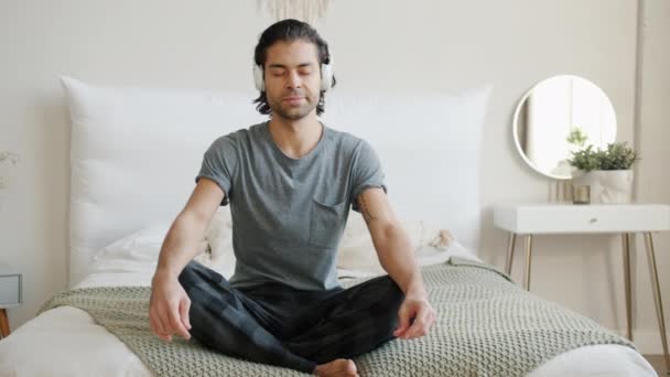 Powolny ruch przystojnego młodzieńca noszącego słuchawki siedzącego w pozycji lotosu w łóżku — Wideo stockowe