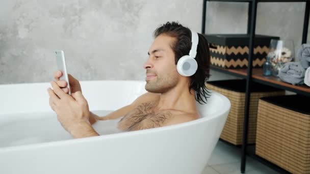 Ragazzo che indossa le cuffie usando lo smartphone e ascoltando musica nella vasca da bagno a casa — Video Stock