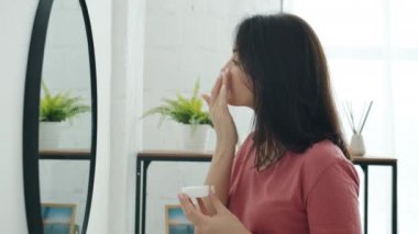 Güzel genç bir kadının banyodaki aynaya bakarak cildine yüz kremi sürmesi.