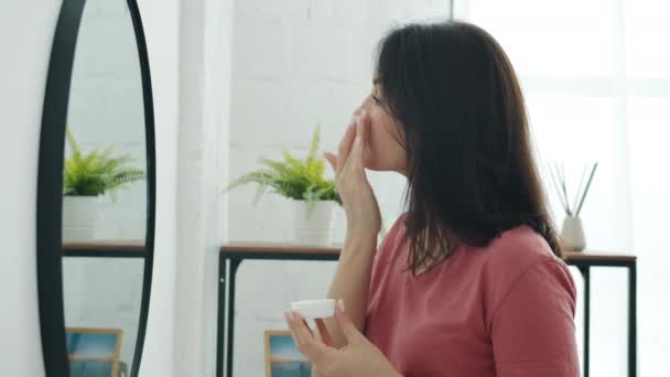Side view van mooie jonge vrouw aanbrengen gezichtscrème op de huid op zoek naar spiegel in de badkamer — Stockvideo