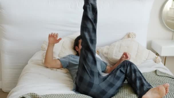 Slow motion of carefree guy in pajamas jumping in bed resting enjoying relaxing leisure time at home — Stock Video