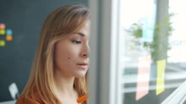 Yaratıcı ofisteki cam tahtaya renkli yapışkan notlarla çalışan genç bir kadın.