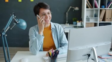 Neşeli ofis çalışanı cep telefonuyla konuşuyor ve bilgisayar ekranının çalışmasına bakıyor.