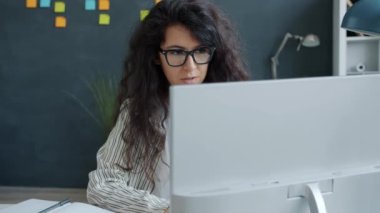 İşten zevk alan genç bir kadın bilgisayar ekranına bakıp el çırpıyor ve ofiste gülümsüyor.