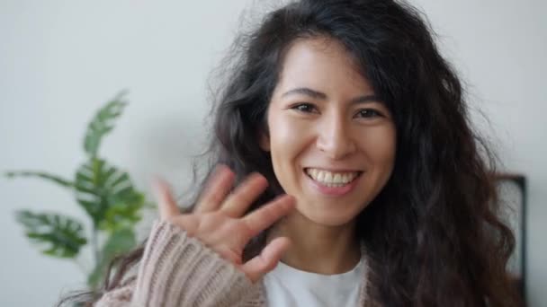 Iloisen sekarotuisen naisen hidas liike hymyilee ja heiluttaa kättään katsoen kameraa kotona — kuvapankkivideo