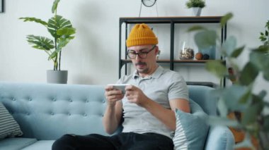 Duygusal genç adam evdeki kanepede akıllı telefon dokunuşuyla video oyunu oynuyor.