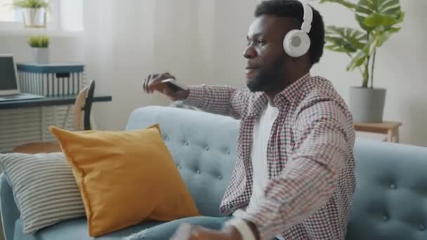 Glad afroamerikansk man som har kul med att lyssna på musik med hörlurar som dansar hemma — Stockvideo