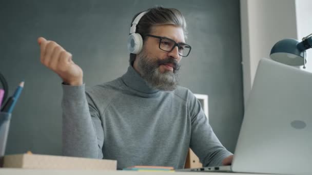 Ώριμος επιχειρηματίας ακούγοντας μουσική μέσω ακουστικών τραγουδώντας και δουλεύοντας με laptop στο γραφείο — Αρχείο Βίντεο