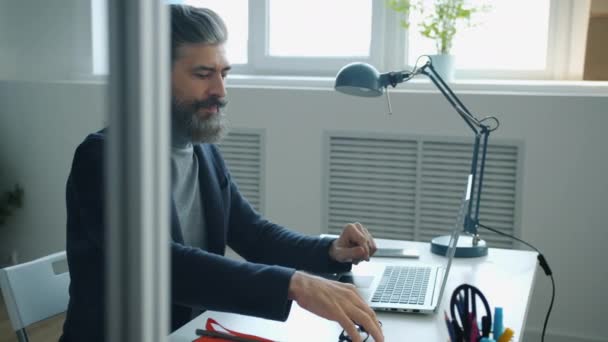 Dojrzały biznesmen noszący okulary za pomocą komputera w pracy, piszący na maszynie przy biurku — Wideo stockowe