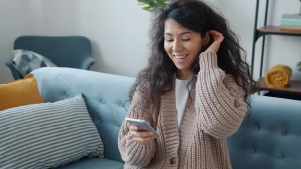 Хрупкая девушка с сенсорным экраном смартфона улыбается, получая хорошие новости, показывая пальцы вверх дома — стоковое видео