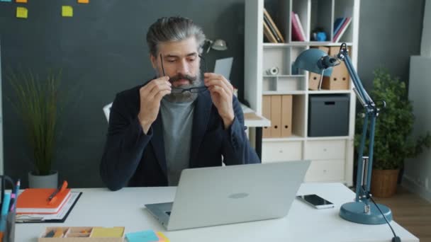 Zwolniony ruch poważnego dojrzałego mężczyzny zakładającego okulary i pracującego z laptopem w biurze — Wideo stockowe