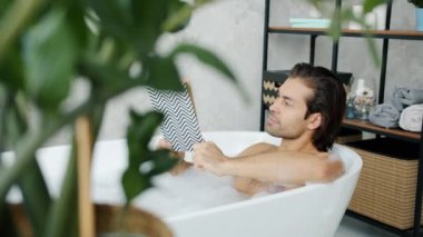 Akıllı adam küvette uzanmış kitap okuyor. Hafif banyoda köpükle sıcak suda dinleniyor.