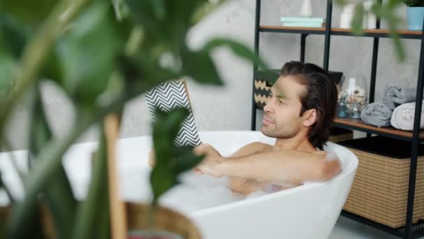 Homem inteligente leitura livro deitado na banheira relaxante em água morna com espuma no banheiro leve — Vídeo de Stock