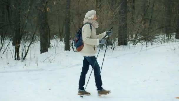 Vista lateral de una anciana caminando en el bosque con bastones de esquí ocupados con deportes de invierno al aire libre — Vídeo de stock